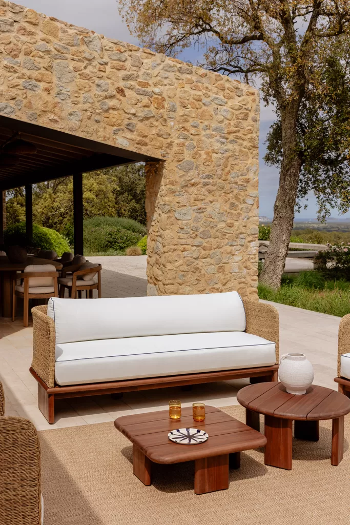 Area relax con divano in fibre naturali e tavolini in legno in un incantevole giardino rustico, sotto un portico in pietra e immersa nella vegetazione mediterranea.