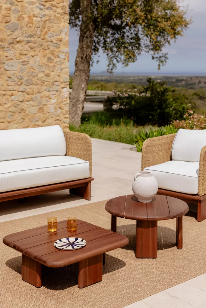 Area relax in un accogliente giardino rustico, con divani in fibre naturali, tavoli in legno massiccio e uno sfondo paesaggistico mediterraneo.