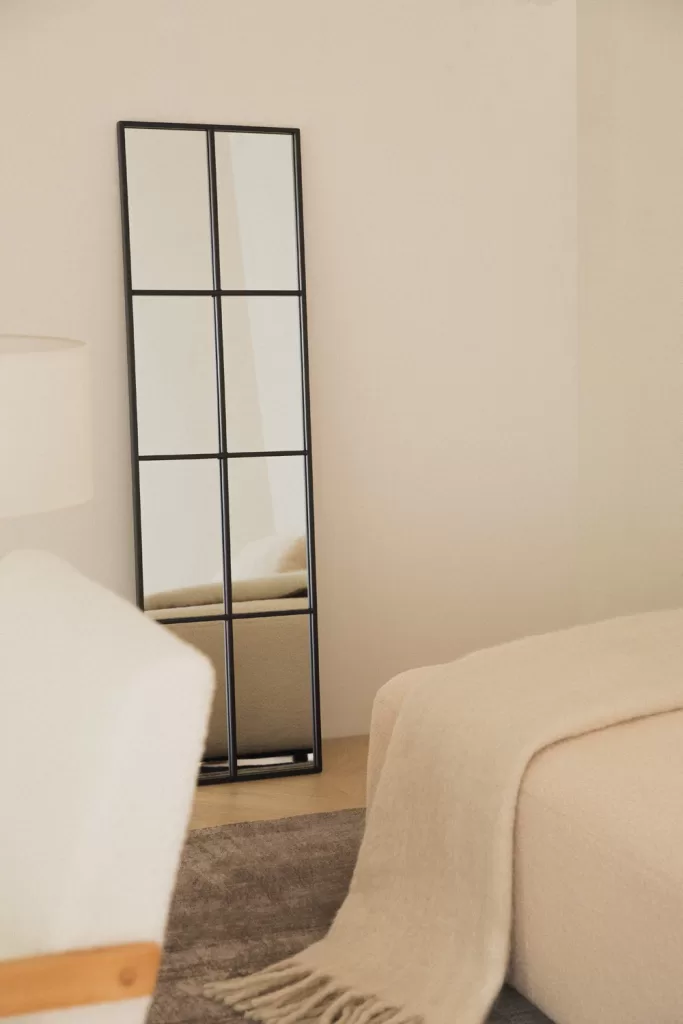 Miroir de sol à cadre en métal noir et couverture au-dessus d'un lit beige dans une chambre grise avec du bois ; touche moderne subtile