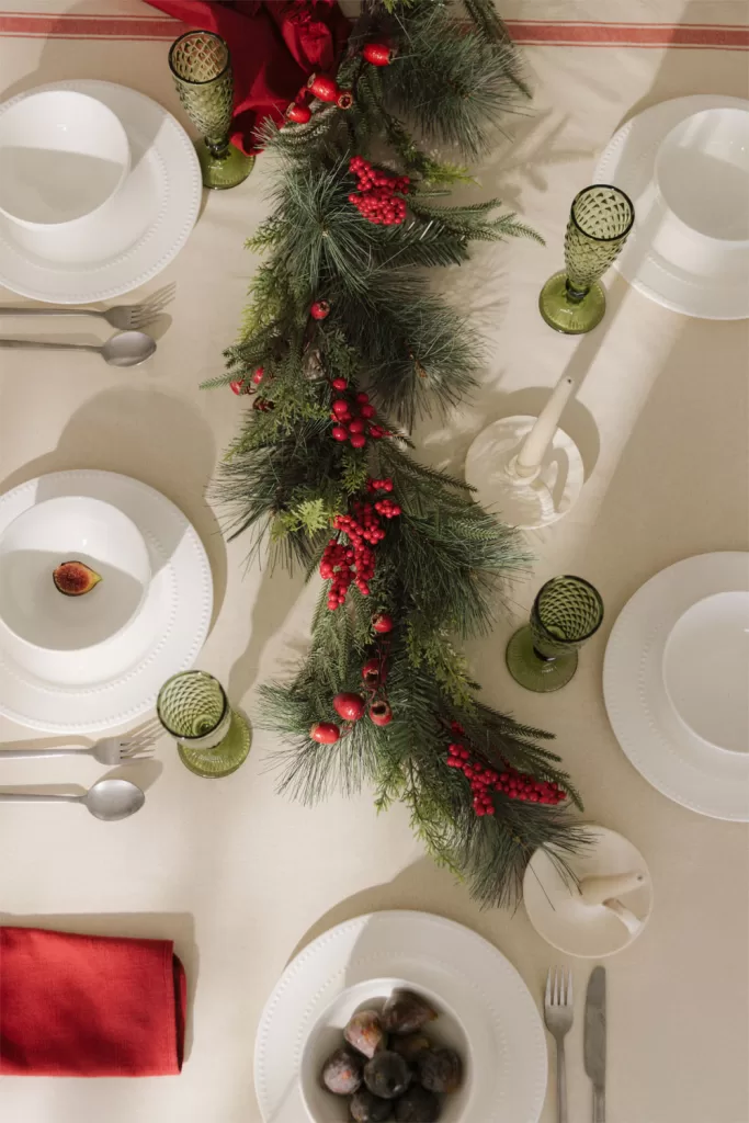 Mesa de jantar decorada com uma grinalda verde e loiça branca, ideal para uma decoração de Natal elegante numa sala pequena.