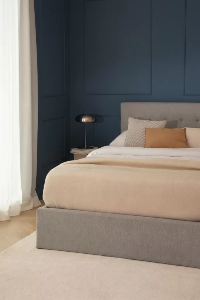 Quarto azul e cinzento com parede azul-marinho escura e cama com sommier cinzento, têxteis bege e almofadas neutras.