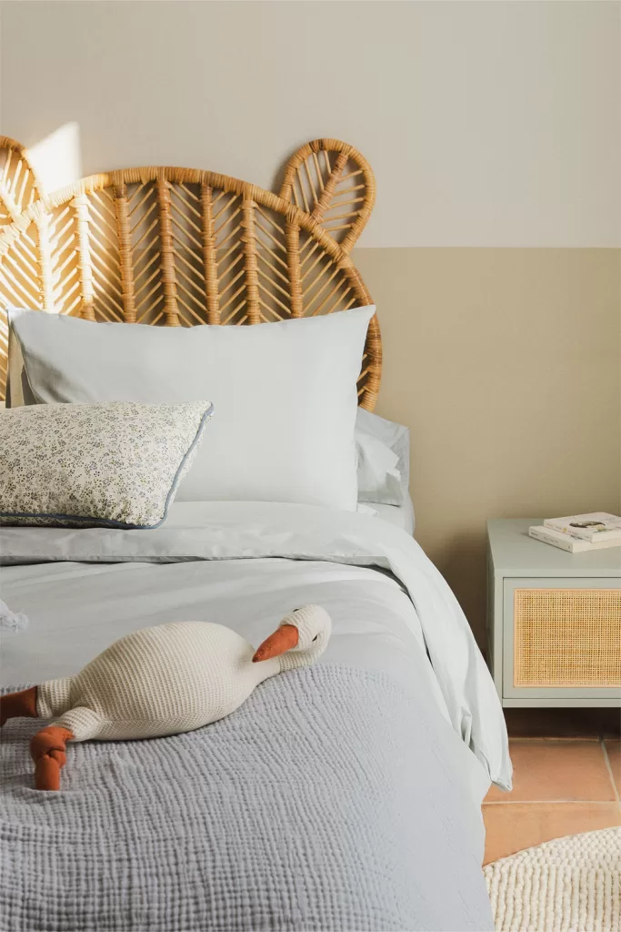 Quarto azul e cinzento com cabeceira em rotim natural, lençóis cinzentos e manta azul, estilo descontraído e luminoso.