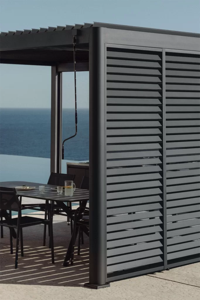 Pérgola bioclimática com painel lateral em alumínio cinza a proteger a mesa e cadeiras no terraço.