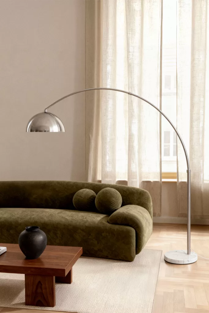 Candeeiro de pé em arco cromado ao lado de um sofá verde e uma mesa de madeira, ideal para iluminar uma sala sem teto falso.